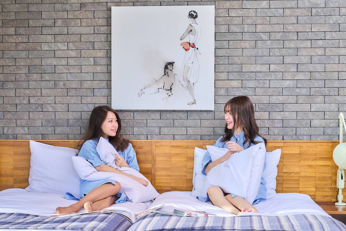 Two people sitting on a bed with a brick wall and a painting in the background.