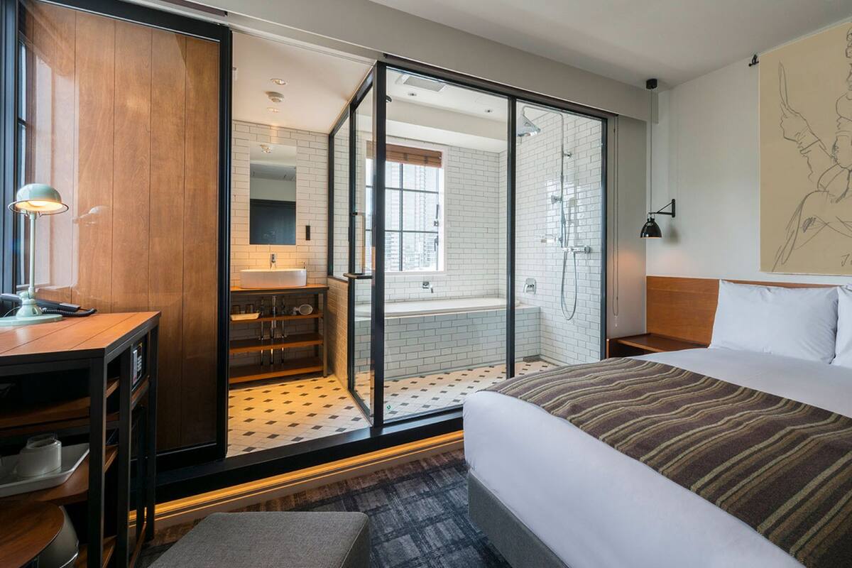 A hotel room with a bed, a desk, a bathroom with a shower, and a large window.