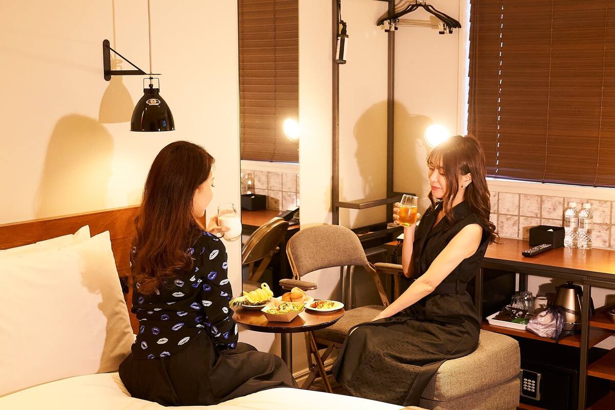 Two women sitting on a bed in a hotel room with a dining table and a kitchenette.