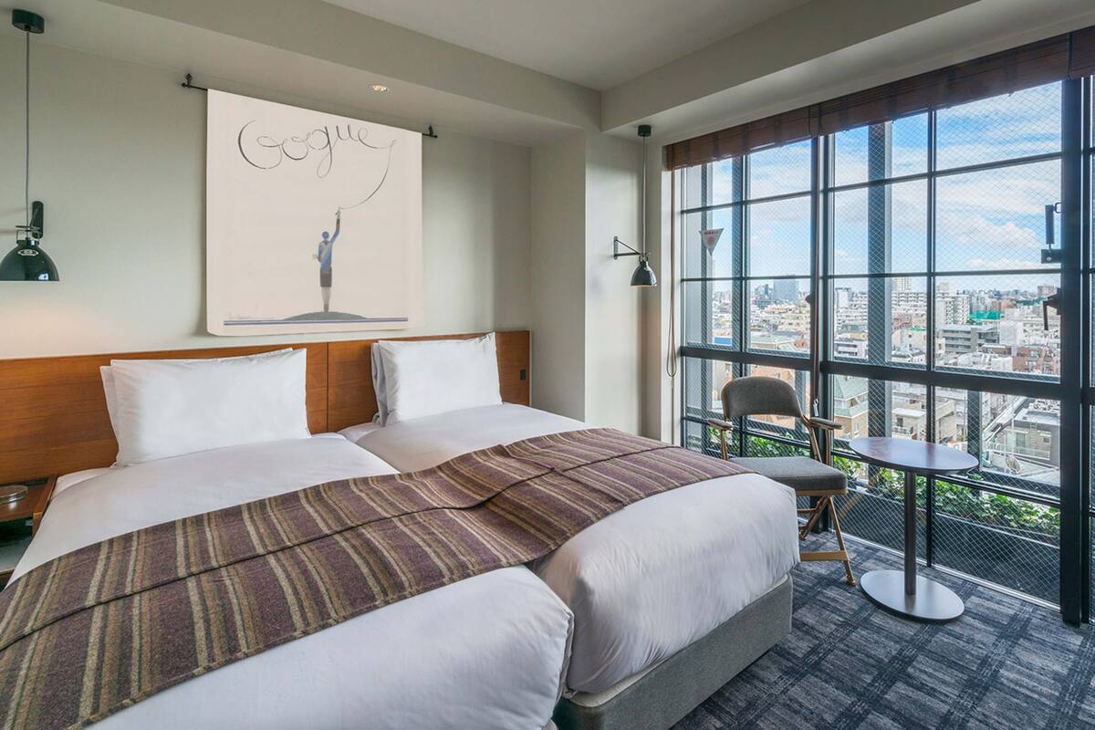 A hotel room with a large bed, a city view through the window, a small table, and a chair.