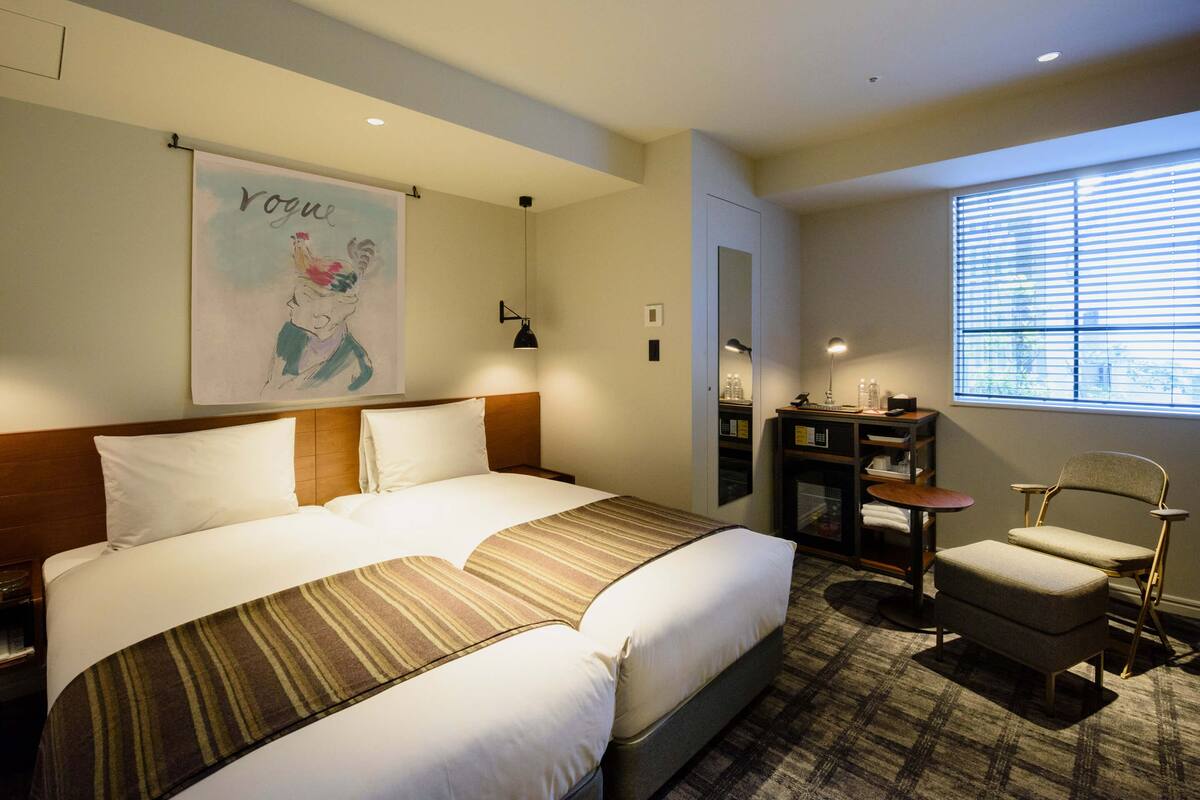 A hotel room with a large bed, a chair, a desk with a lamp, a mirror, and a window with blinds.
