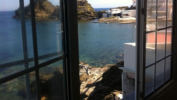 A coastal view through a window with a clear view of the sea, rocky terrain, and buildings on the shore. - LIKE BEING ON BOARD OF A BOAT AT SEA (MAHON)