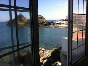 Vista costera a través de una ventana, con mar despejado, terreno rocoso y edificios en la orilla.