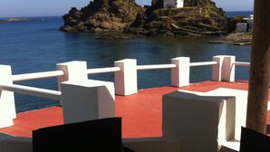 A terrace with white chairs and tables overlooking a rocky coastline with a small white building on a rock formation. - LIKE BEING ON BOARD OF A BOAT AT SEA (MAHON)