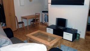 Un espace de vie compact comprenant une table à manger en bois, un canapé, une table basse avec un plateau en verre, un téléviseur posé sur un meuble noir et une armoire en bois.