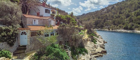 A stone house with a tiled roof, located on a cliff overlooking a clear blue sea.