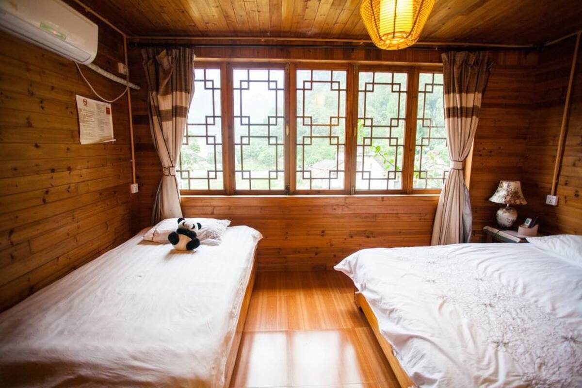 A wooden cabin room with two beds, a window with a geometric pattern, and a hanging light fixture.