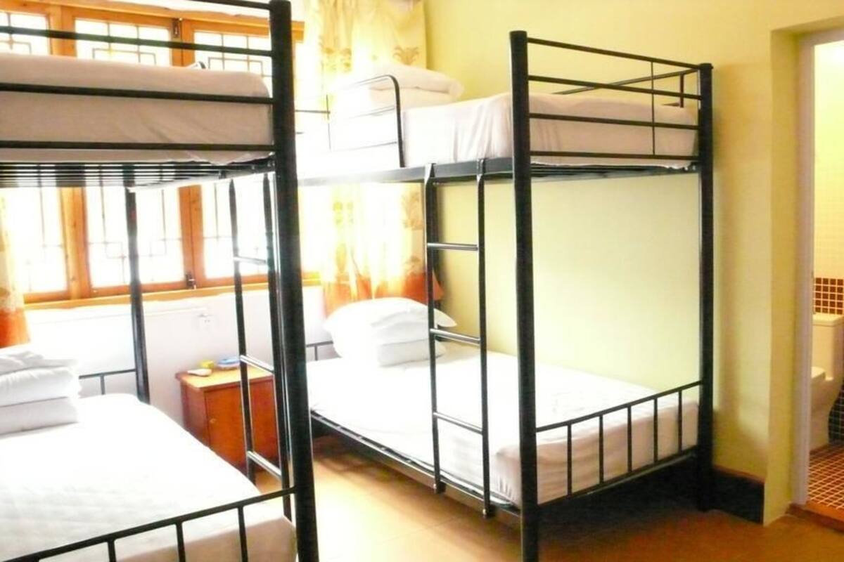 A dormitory room with bunk beds, a small bedside table, and a window with curtains.
