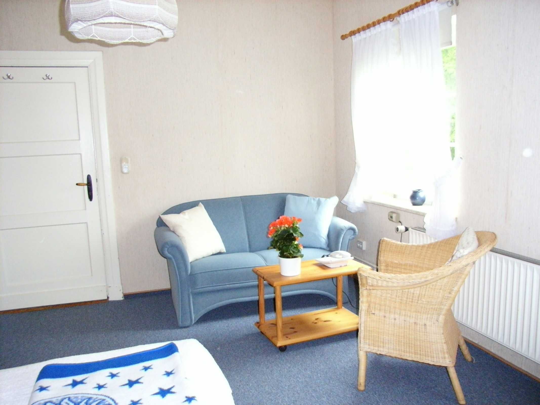 Une pièce avec un canapé bleu, un fauteuil en osier, une petite table en bois, une porte blanche et une fenêtre avec des rideaux.