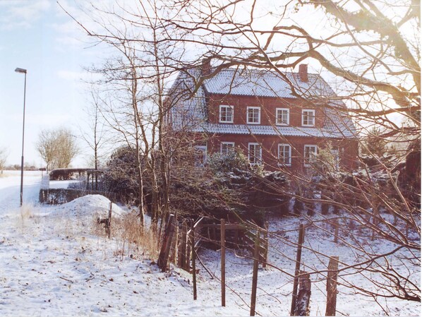 Paisagem coberta de neve com uma casa de tijolos, árvores sem folhas e uma cerca.