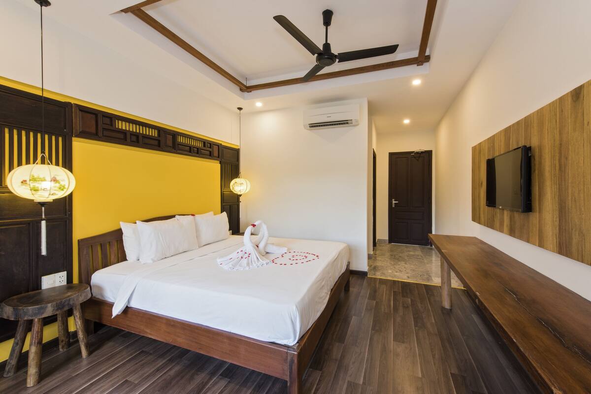 A hotel room with a wooden bed, a ceiling fan, a TV mounted on the wall, and a bench.