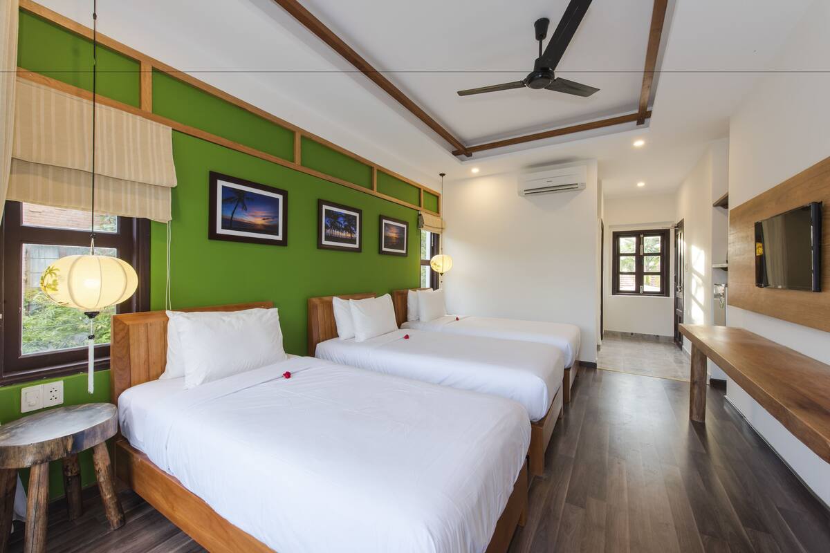 A hotel room with three single beds, a wooden bench, a ceiling fan, and a TV mounted on the wall.