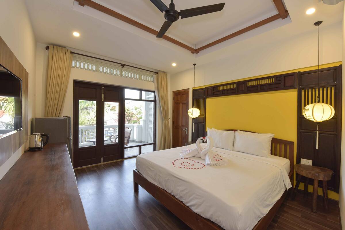 A hotel room with a large bed, a ceiling fan, a balcony with a view, and a wooden floor.