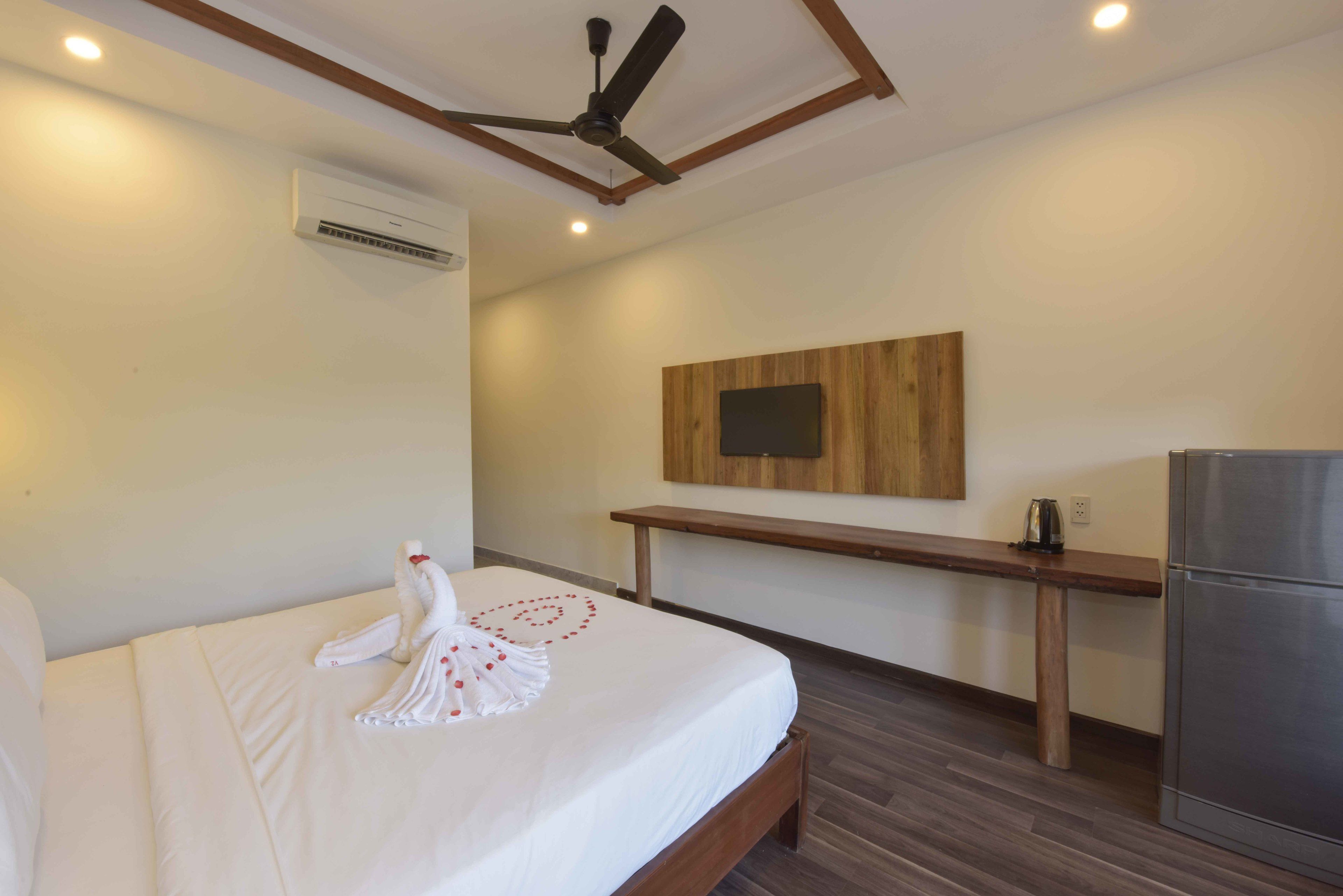 Une chambre d’hôtel équipée d’un lit, d’un ventilateur de plafond, d’un banc en bois, d’un téléviseur fixé au mur et d’un réfrigérateur.