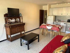 A compact living space with a dining table, chairs, a sofa, a television on a wooden cabinet, and a kitchenette.