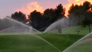 Un campo de golf con múltiples aspersores en el green, árboles al fondo y una bandera señalando un hoyo.