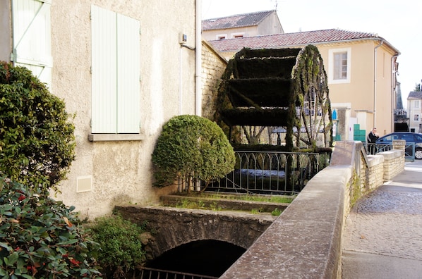 Un pont de pierre avec une petite arche, un bâtiment blanc avec des volets et un arbre dont les branches sont recouvertes de mousse.