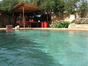 A clear pool with a wooden pergola and seating area, surrounded by greenery and stone walls.