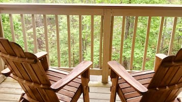Two wooden Adirondack chairs on a wooden deck with a white railing, overlooking a forested area.