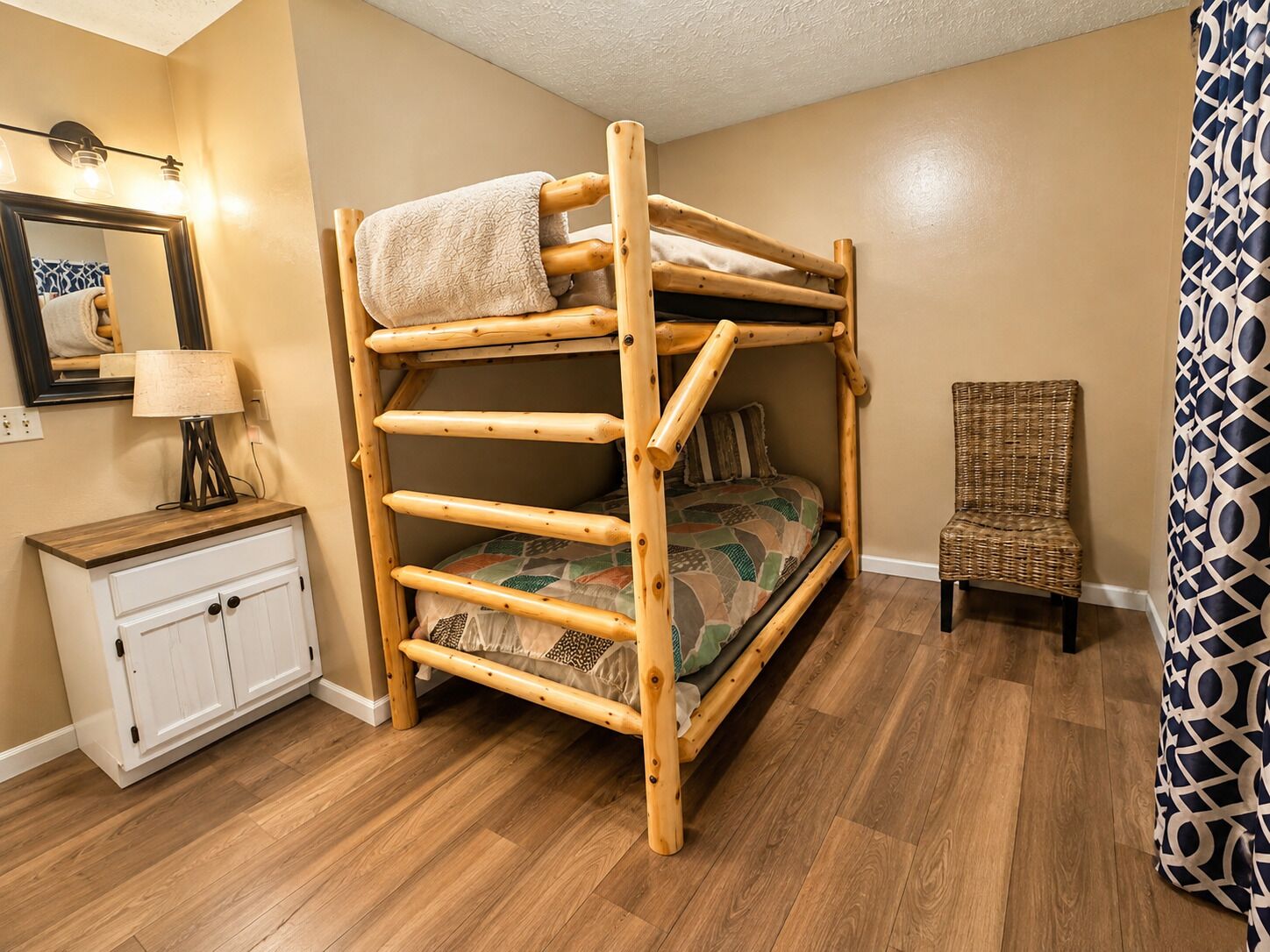 A bunk bed with a wooden frame, a bedside table with a lamp, a chair, and a patterned curtain.