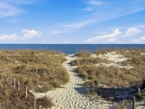Un sentiero sabbioso che conduce verso l'oceano, circondato da erba secca e pali di legno.