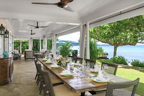 A covered outdoor dining area with a view of the sea, furnished with wicker chairs and a long wooden table set for a meal. - Beachfront Villa, Staff, Boat, Pool, Tennis, Gated (Montego Bay)