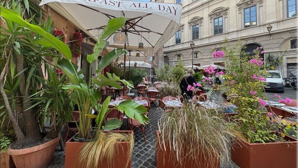 Un café al aire libre con un letrero que dice "BREAKFAST ALL DAY" y plantas en macetas alrededor de las mesas.