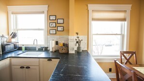 A kitchen with a black marble countertop, white cabinets, and a window with blinds. There are framed pictures on the wall and a microwave on the counter.