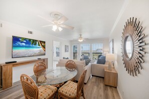 A modern living room with a glass dining table, wicker chairs, a flat-screen TV, and a ceiling fan.