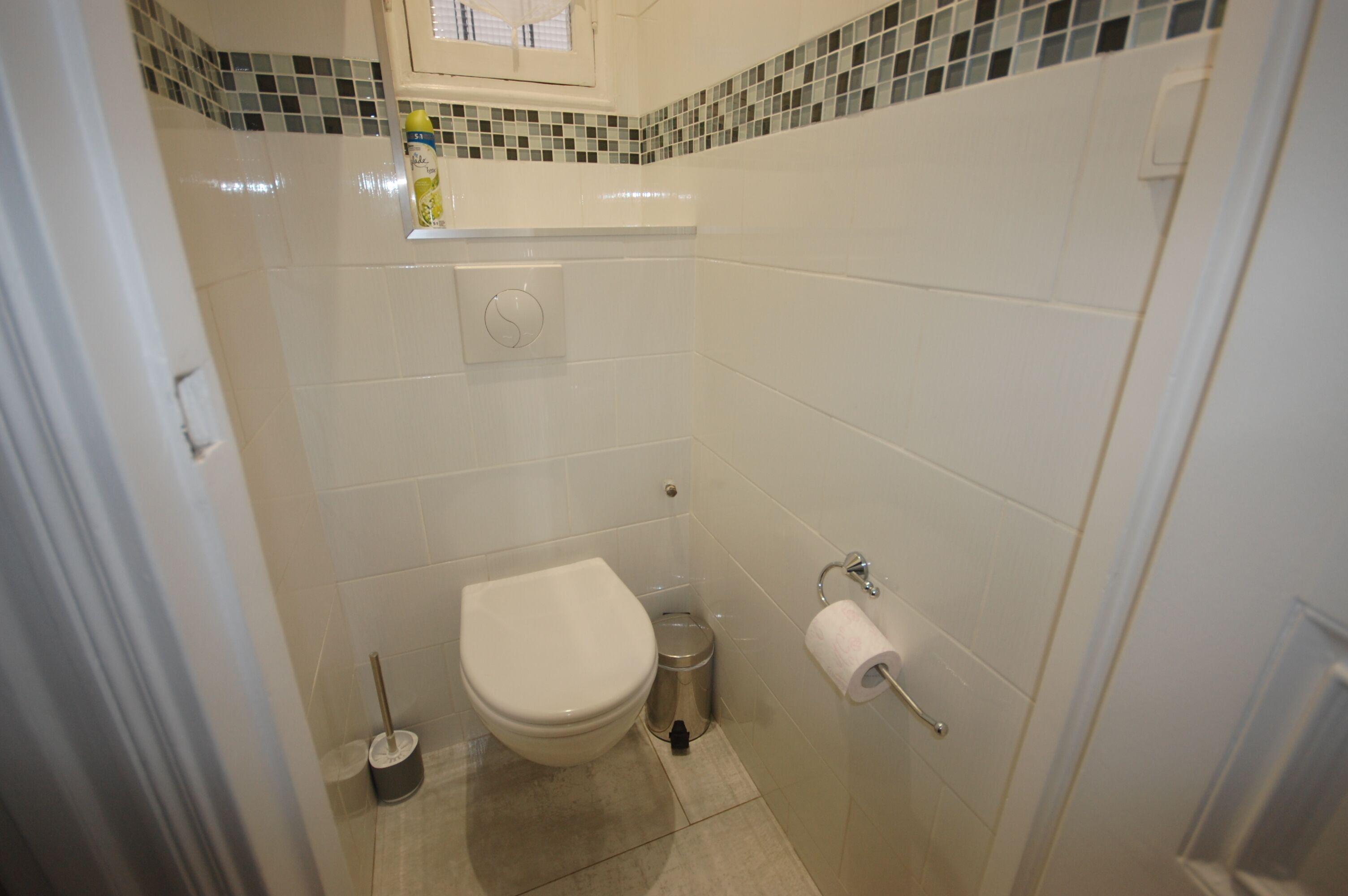 A small bathroom with a toilet, a trash can, a toilet paper holder, and a window above the toilet.
