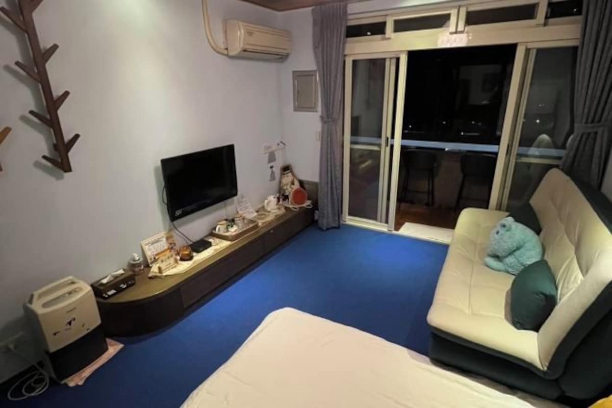 Habitación de hotel con televisión de pantalla plana, un sofá con un oso de peluche, una cama, alfombra azul y una puerta corredera de vidrio.
