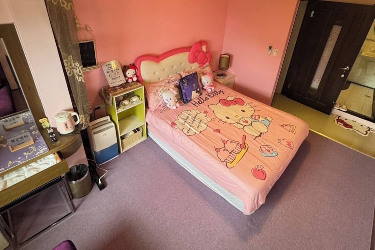 A bedroom with a pink wall, a bed with Hello Kitty bedding, a wooden ceiling, a small table with a kettle, and a door.