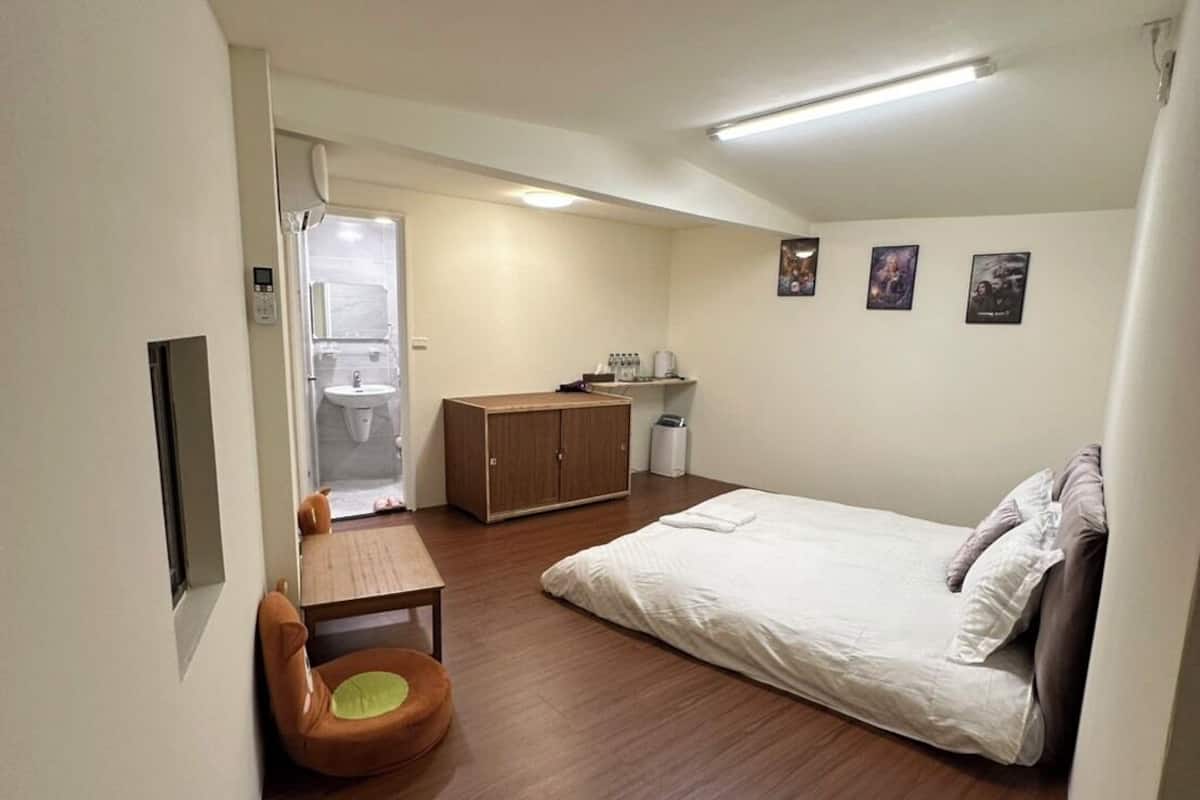 A compact bedroom with a single bed, a small wooden table, a chair, a wardrobe, and a bathroom visible through a glass door.
