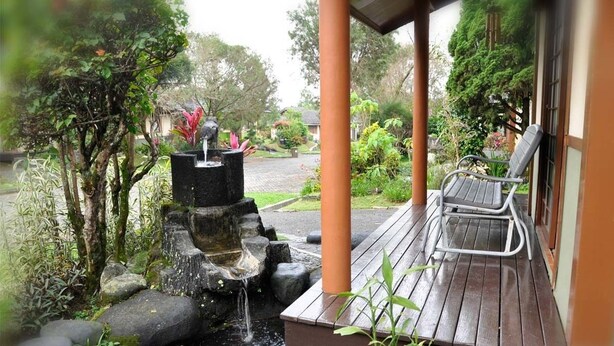 A wooden deck with a bench, a small water feature with a statue, and lush greenery. - Villa Kota Bunga Ade (Cipanas)