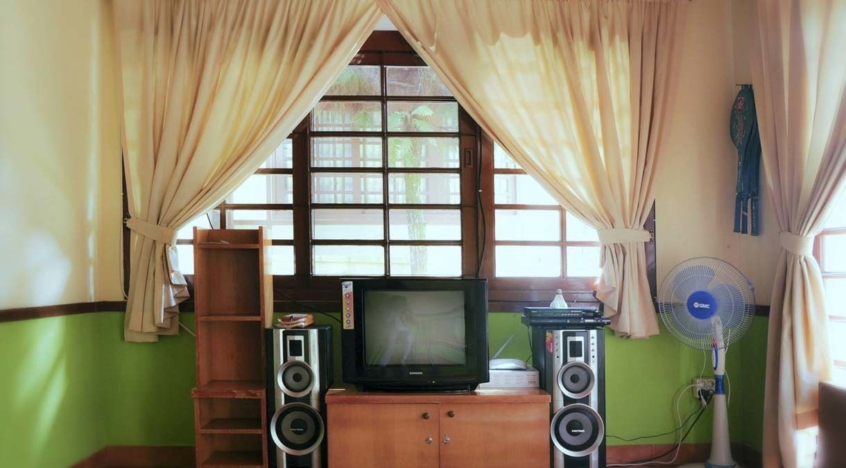 Una habitación con una televisión sobre un mueble de madera, dos altavoces, una ventana con cortinas y un ventilador fijado a la pared.