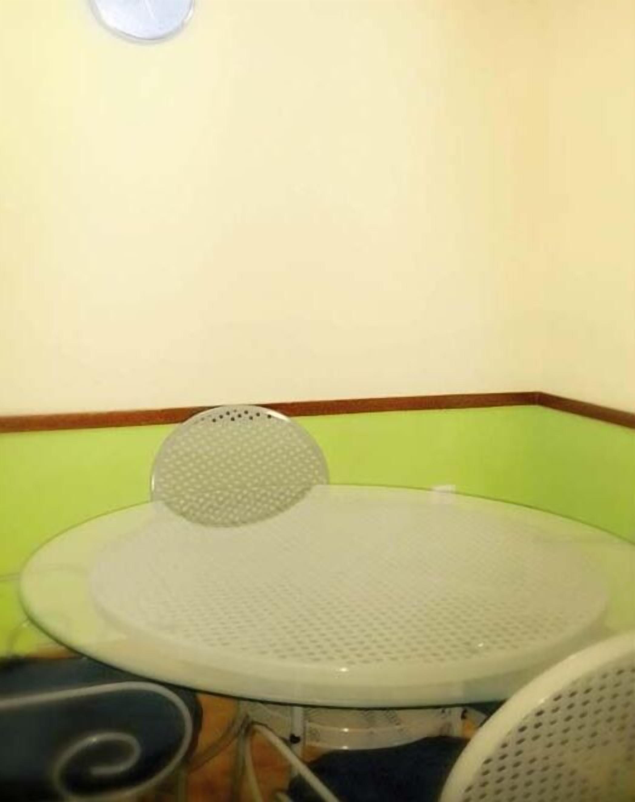 A round glass table with a metal frame, a chair with a perforated backrest, and a green and white wall with a brown trim.