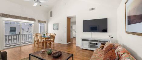 A spacious living room with a large TV, wooden flooring, a dining area with chairs, and a balcony with a railing.