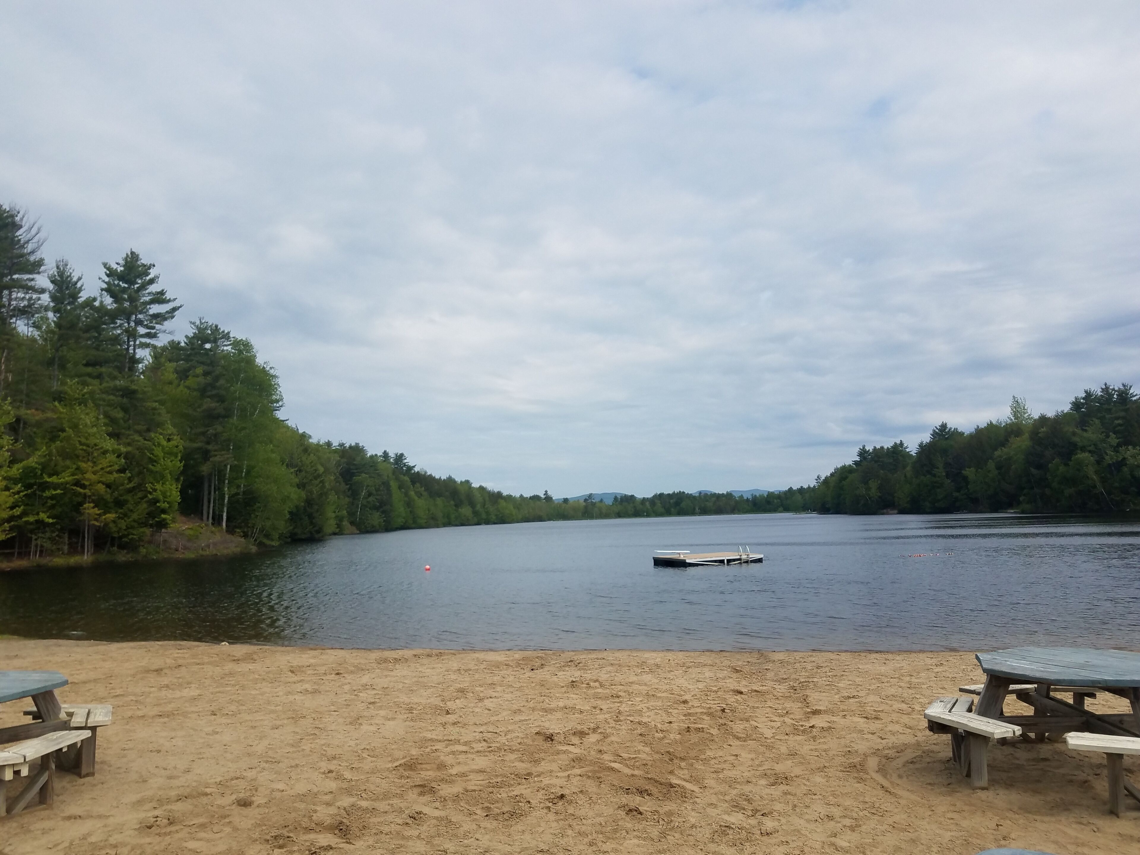 Une vue sur le lac, avec une plage de sable, deux tables de pique-nique et une jetée qui s’avance dans l’eau.