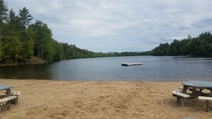 A lakeside view with a sandy beach, two picnic tables, and a dock extending into the water.