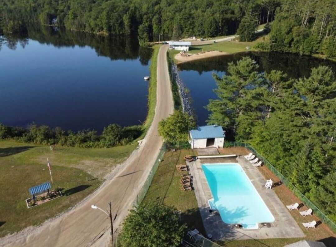 Vue aérienne d’une propriété comprenant une piscine, un petit bâtiment et une jetée s’avançant dans un lac.