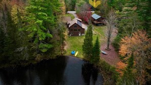 A cabin with a large window, surrounded by dense forest and a lake.