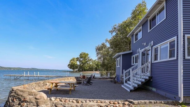 A two-story building with a stone deck and outdoor seating area by a body of water. - Deep Run Fun - Luxury Lake Life (Canandaigua)