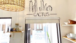 A room with a wall-mounted cactus illustration, a wicker pendant light, and a window with a view of a building outside. - Loreto80- NEW Studio Cactus, in downtown by beach (Loreto)