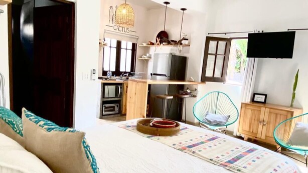 A bedroom with a bed, a bedside table, a chair, and a view of a kitchen area. - Loreto80- NEW Studio Cactus, in downtown by beach (Loreto)