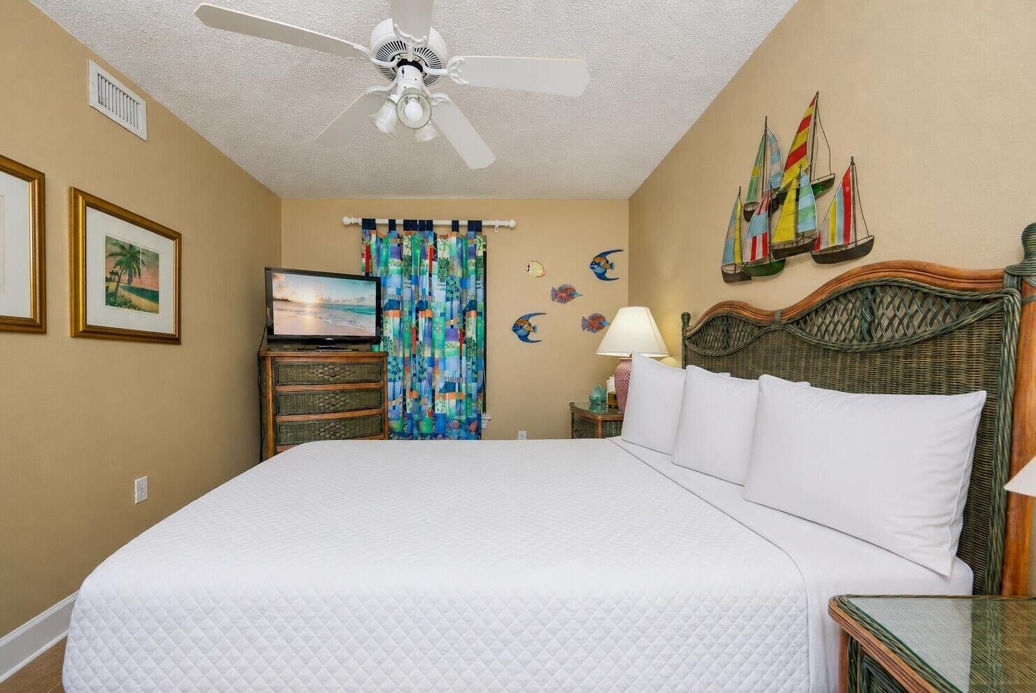 A hotel room with a bed, a ceiling fan, a television, a wall with sailboats, and a window with curtains.