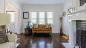 A living room with a brown leather sofa, a white armchair, and a floor lamp. There is a fireplace with a black brick surround and a large window with blinds.