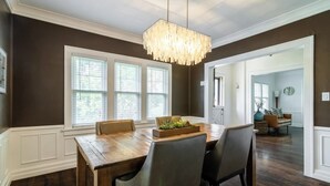 A dining area with a wooden table, four chairs, a chandelier, and large windows.