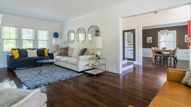 A spacious living room with a dark wood floor, a white sofa with yellow cushions, a coffee table, and a dining area with a wooden table and chairs.
