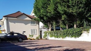 Een oprit met een geparkeerde auto, een huis met een pannendak en een grote boom die schaduw geeft.