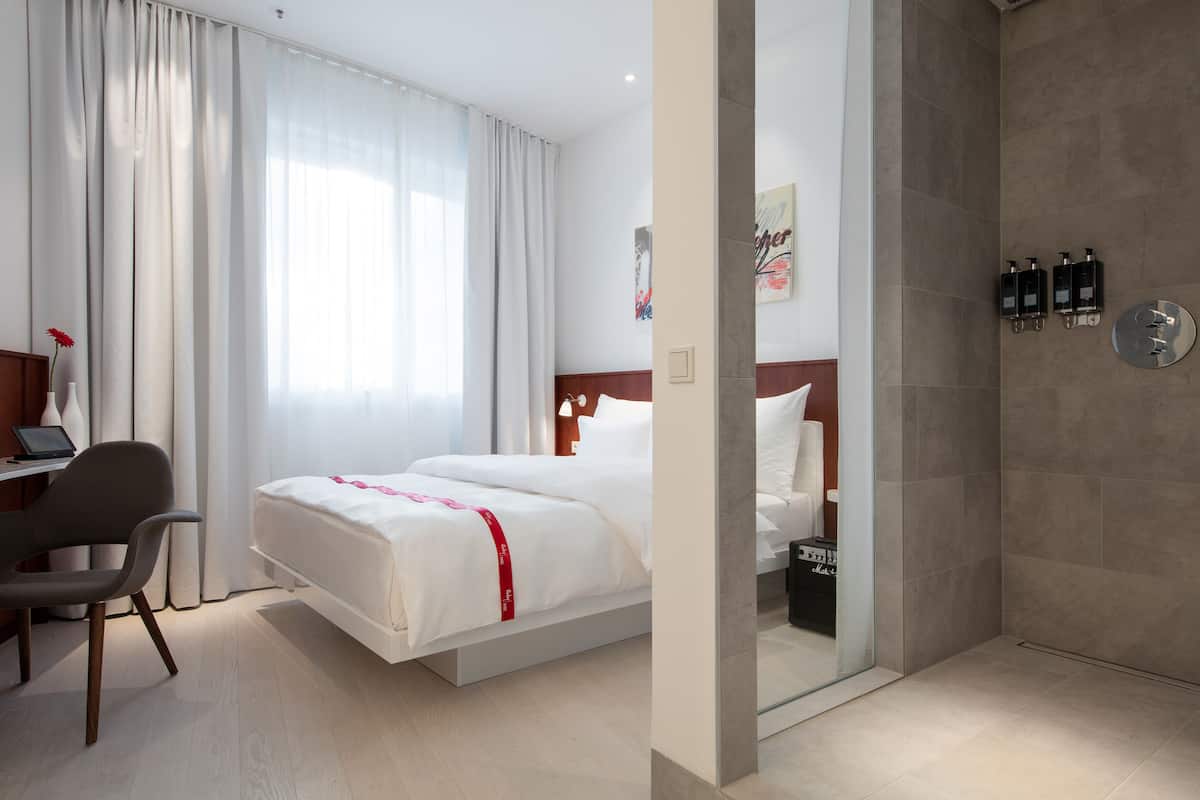 A hotel room with a bed, a desk, a chair, and a shower area.